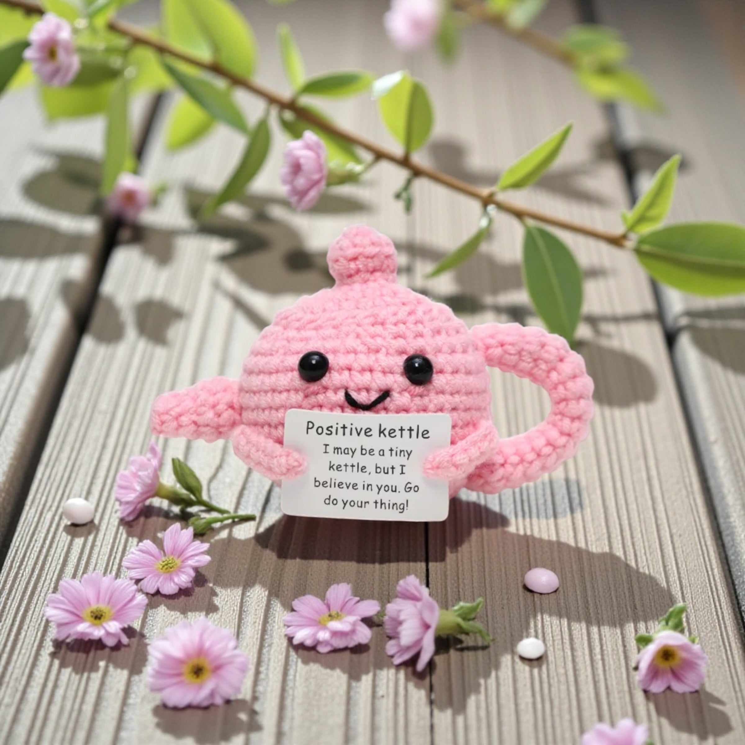 Pink knitted kettle with a motivational tag on a wooden surface with flowers