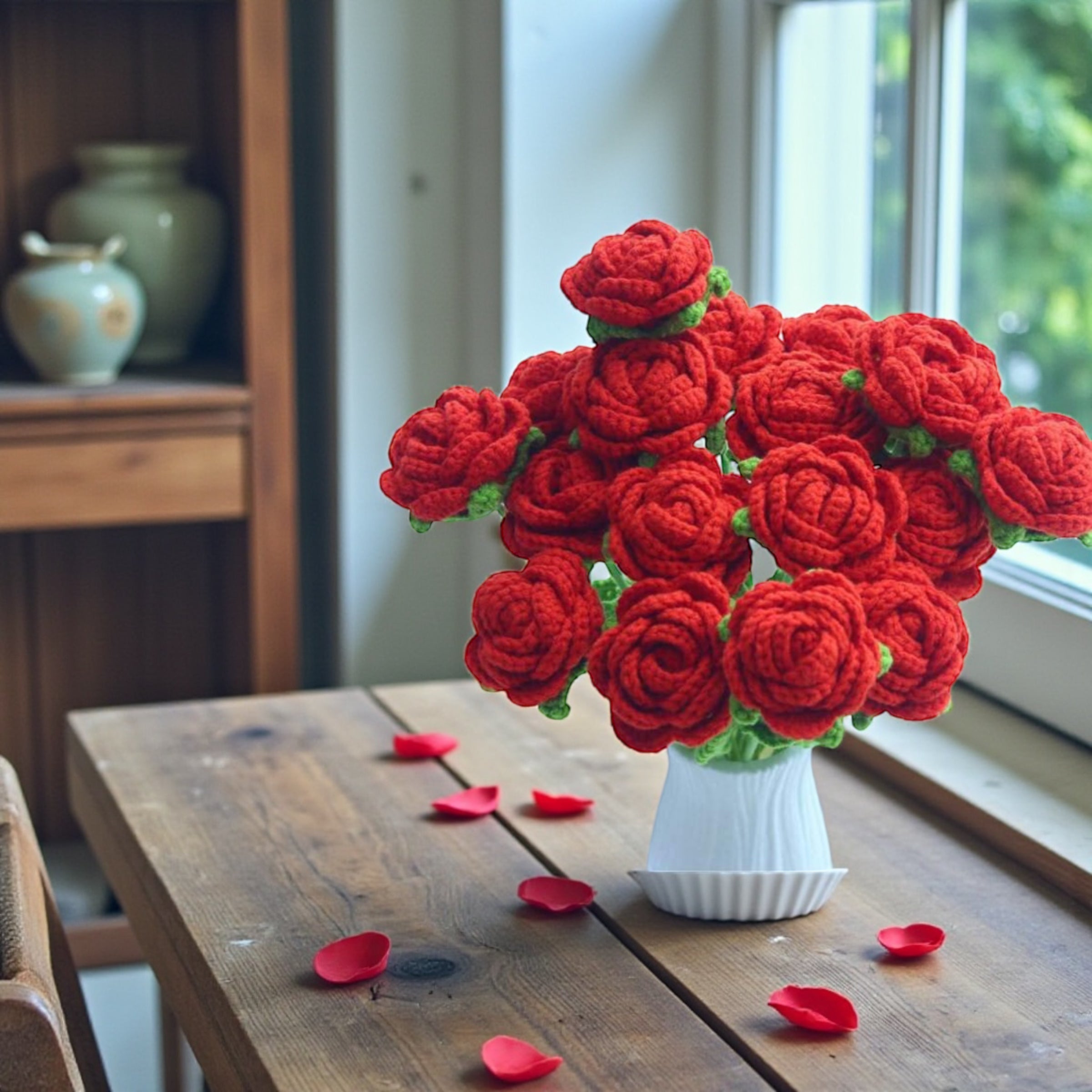 Crocheted red roses in a white vase on a wooden table with scattered rose petals.