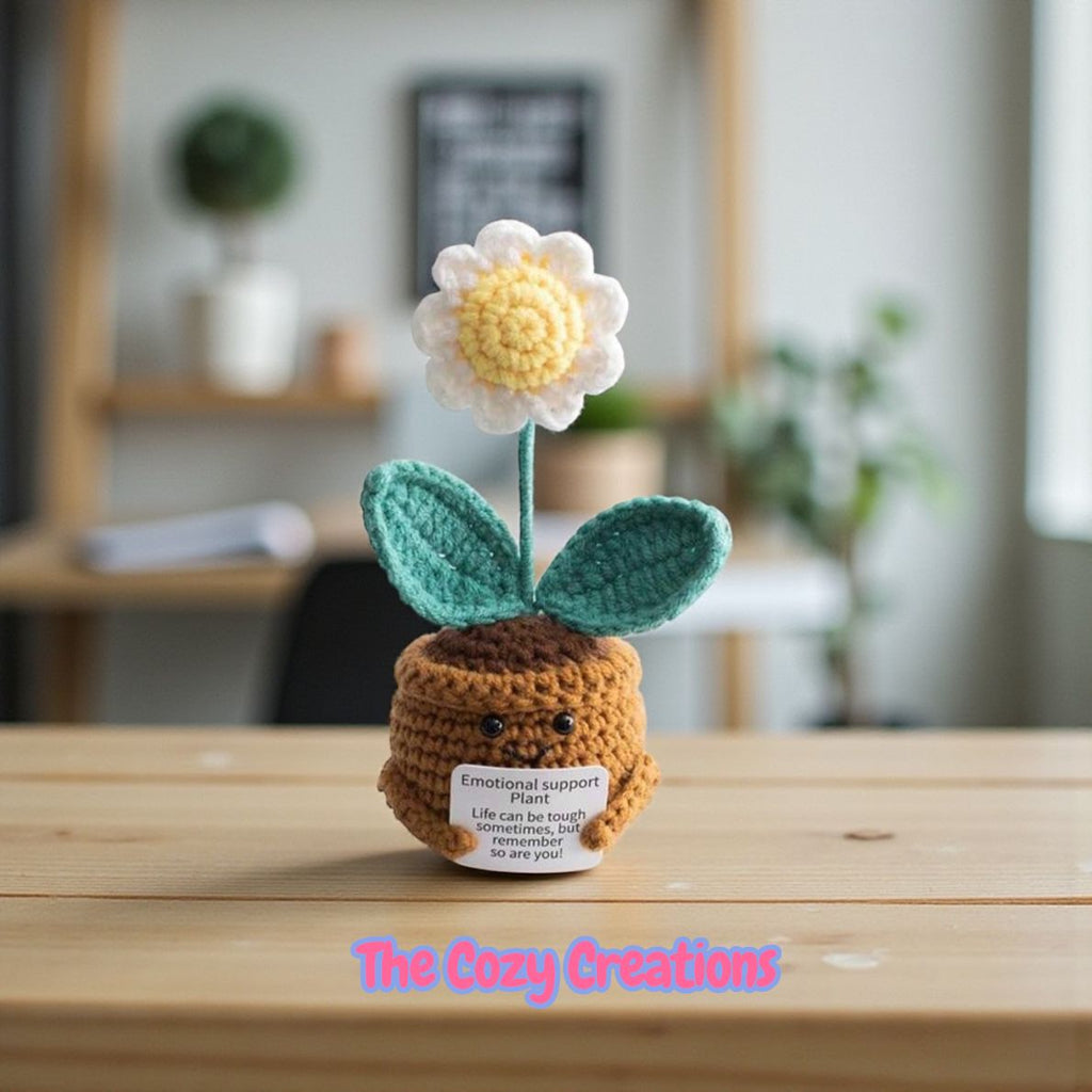 Crocheted flower pot plant with a face on a wooden surface, branded 'The Cozy Creations'.