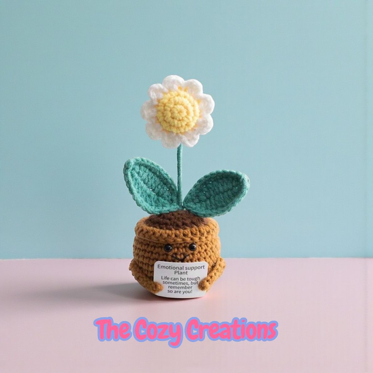 Crocheted flower pot with a flower on a pink surface and light blue background, branded 'The Cozy Creations'.