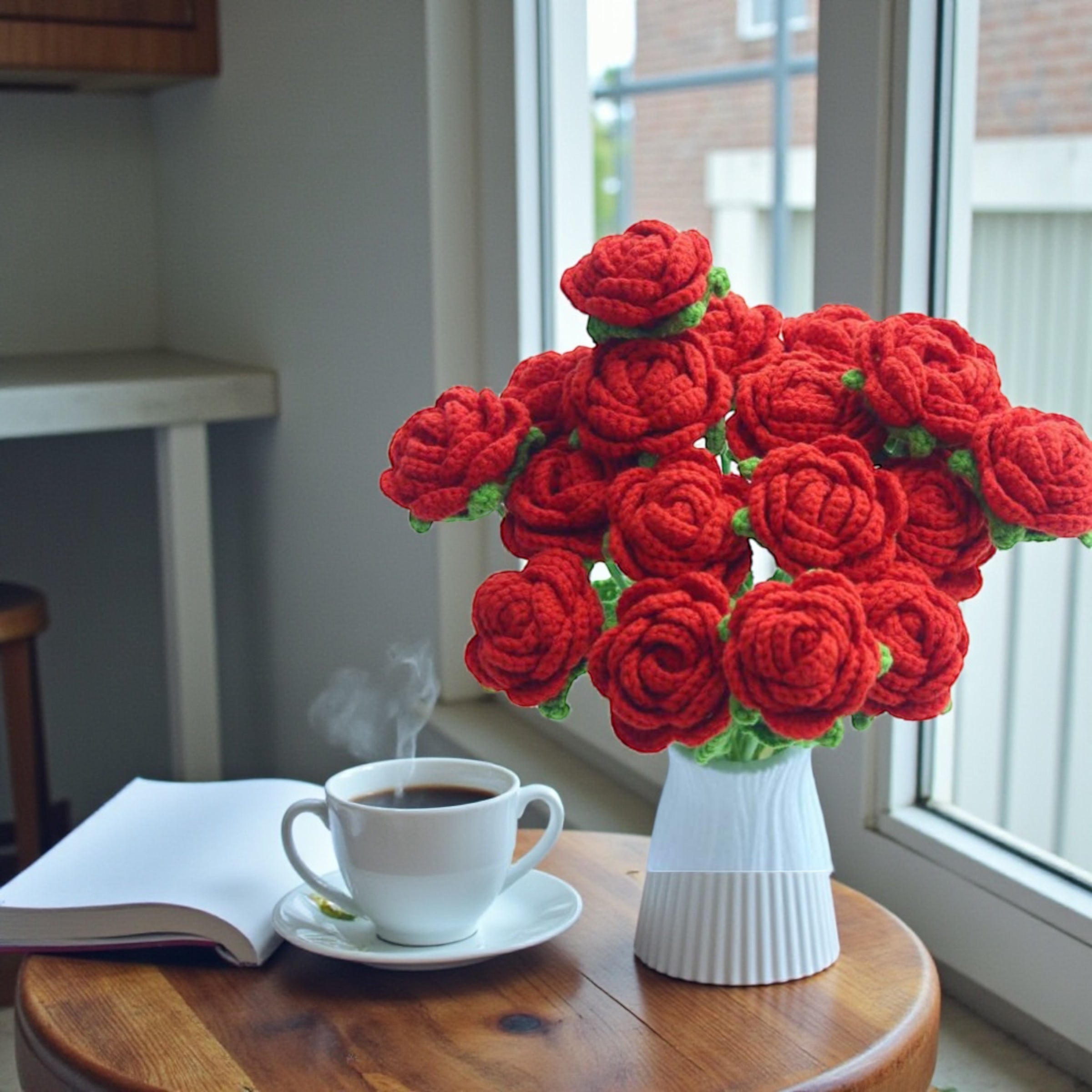 Crocheted red flowers in a white vase on a wooden table with a cup of coffee and an open book.