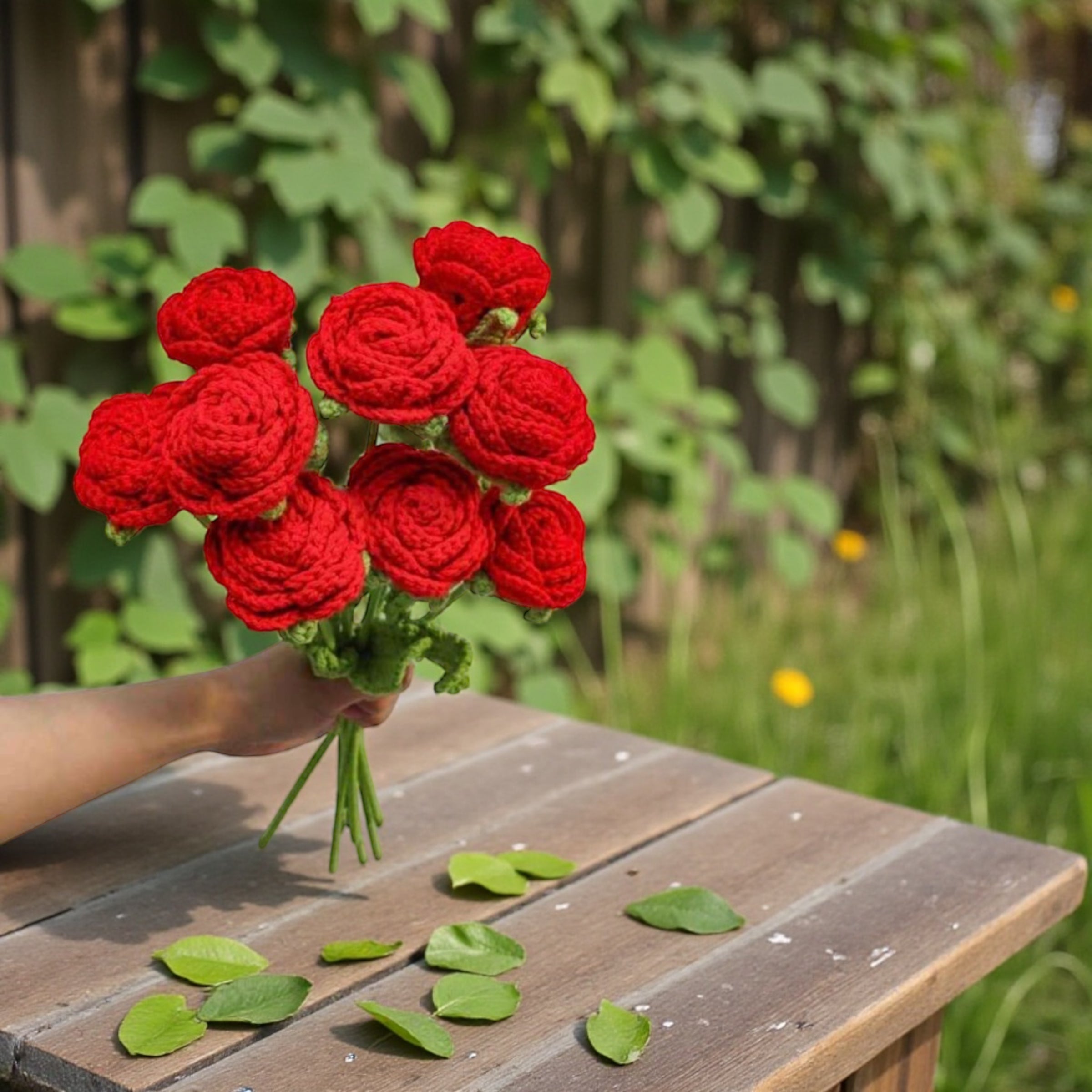 Bouquet of red crocheted roses held by a hand on a wooden table with green leaves.