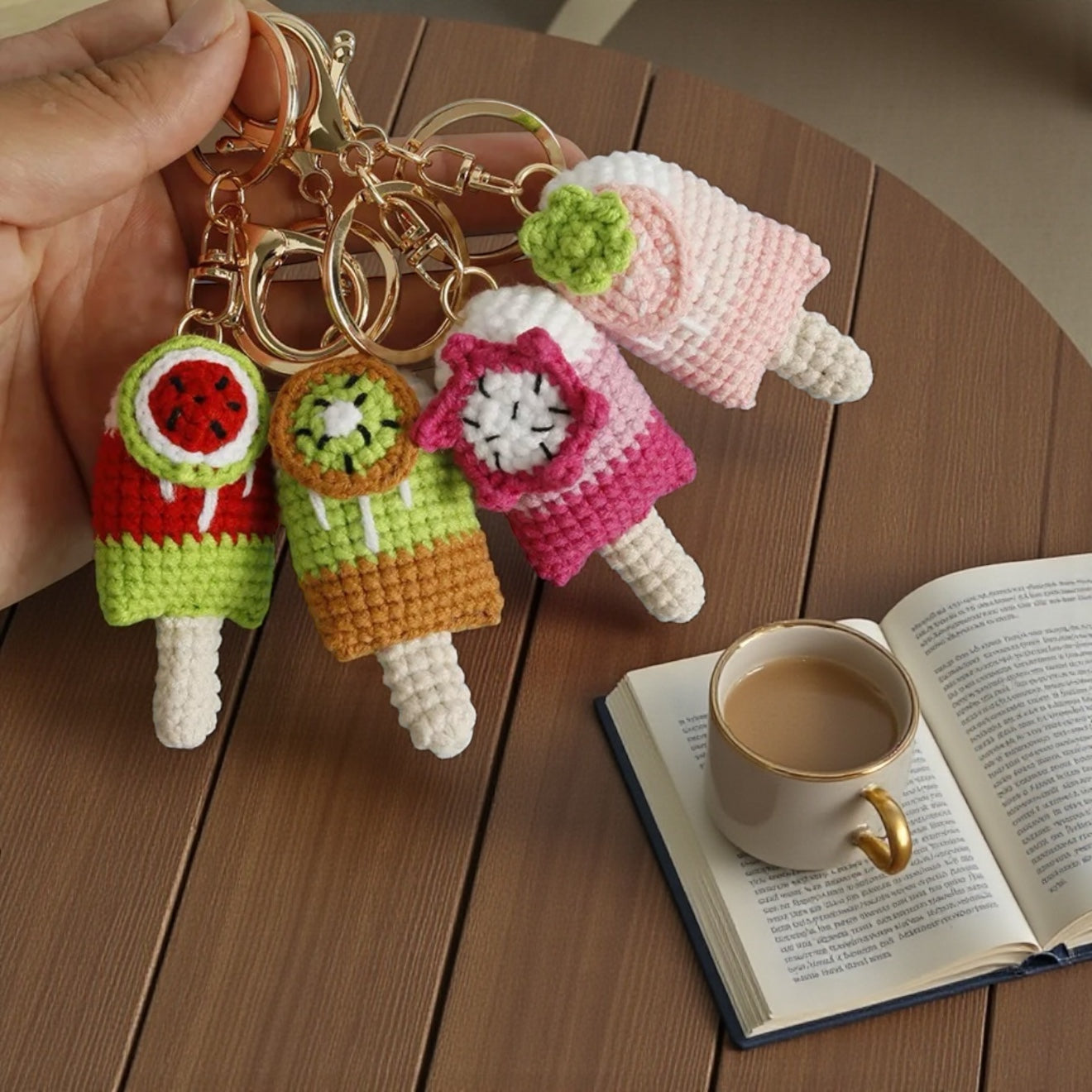 Hand holding a set of colorful crochet keychains on a wooden surface with a book and coffee cup.