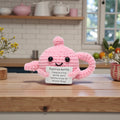 Pink knitted teapot with a motivational message on a wooden surface in a kitchen setting