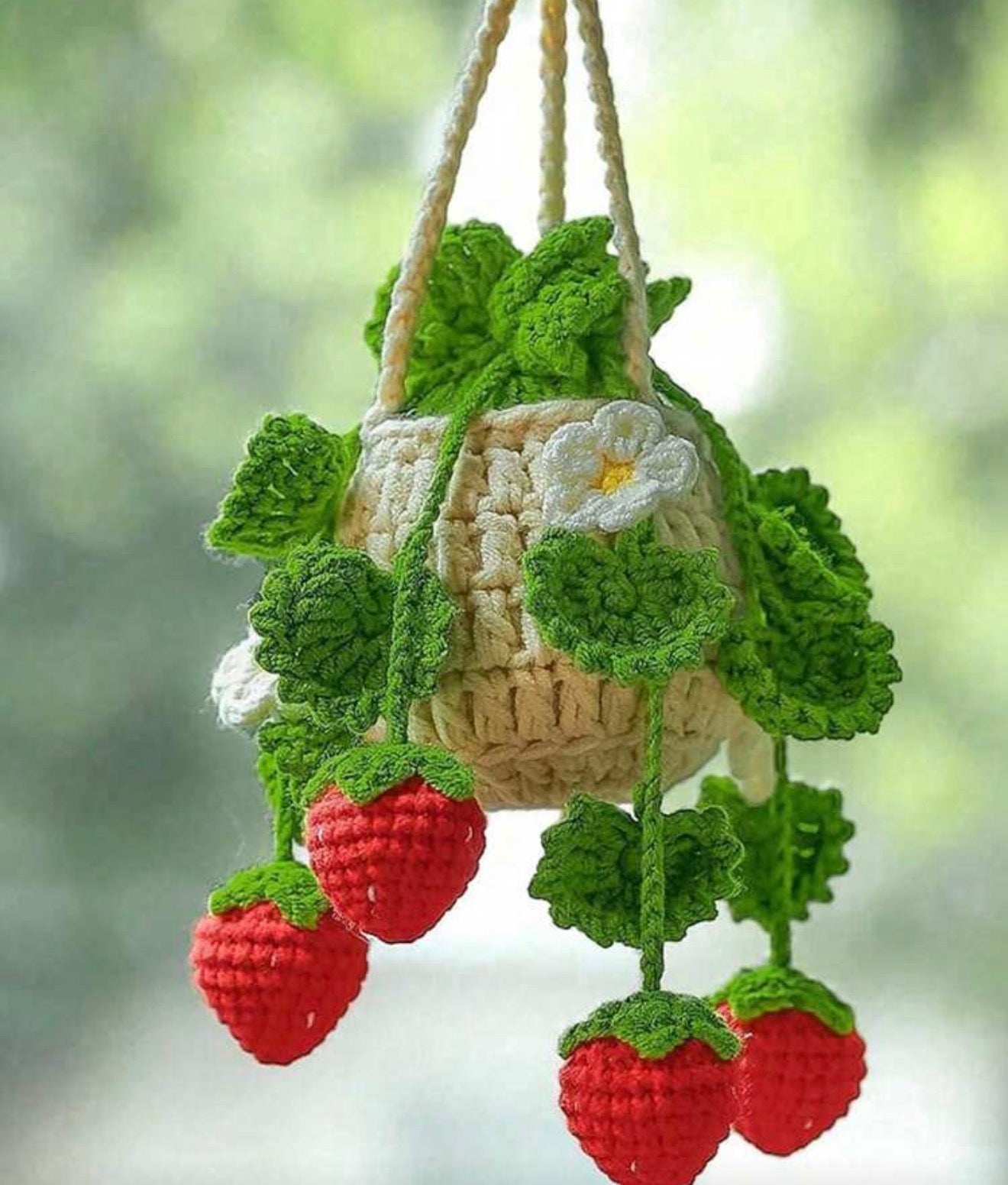 Crocheted strawberry plant with leaves and a flower against a blurred green background