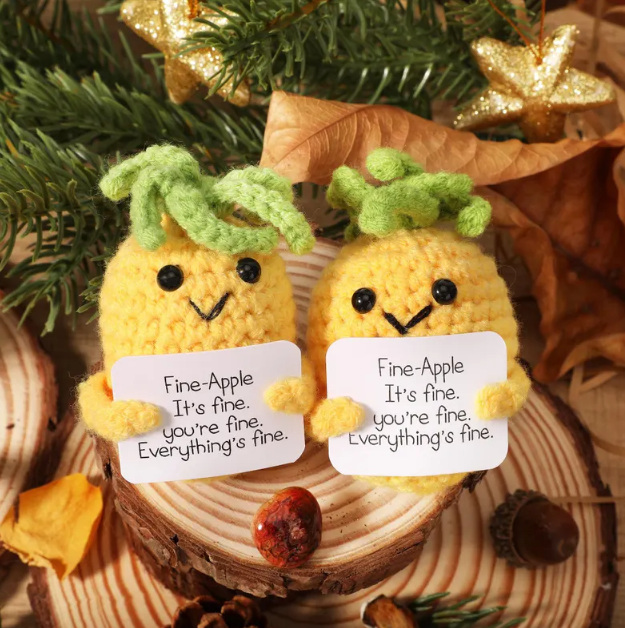 Two knitted pineapple characters holding signs with messages on a wooden surface with autumn leaves and pine branches.
