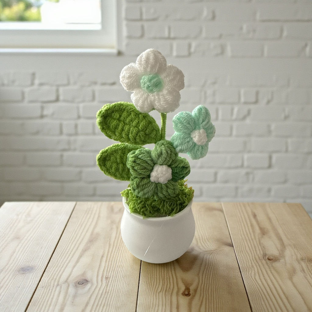 Knitted flower arrangement in a white pot on a wooden surface with a white brick wall background.