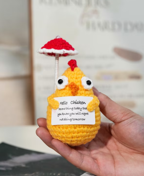 Hand holding a small yellow crocheted chicken with a red umbrella and humorous message.