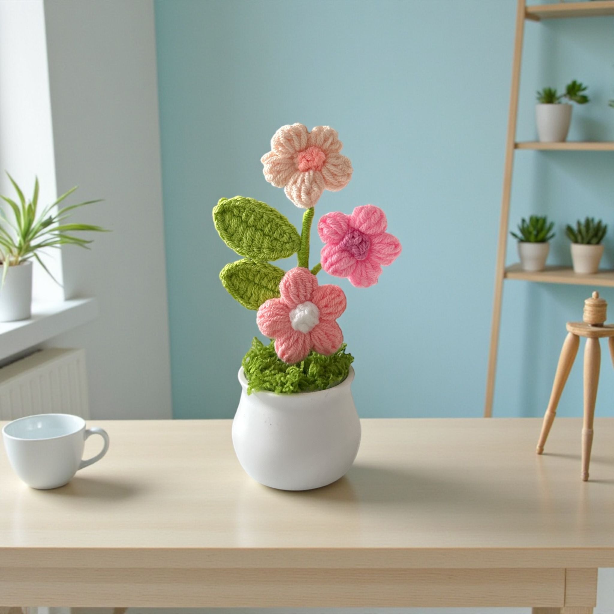 Crocheted flowers in a white pot on a table with a cup, against a light blue wall.