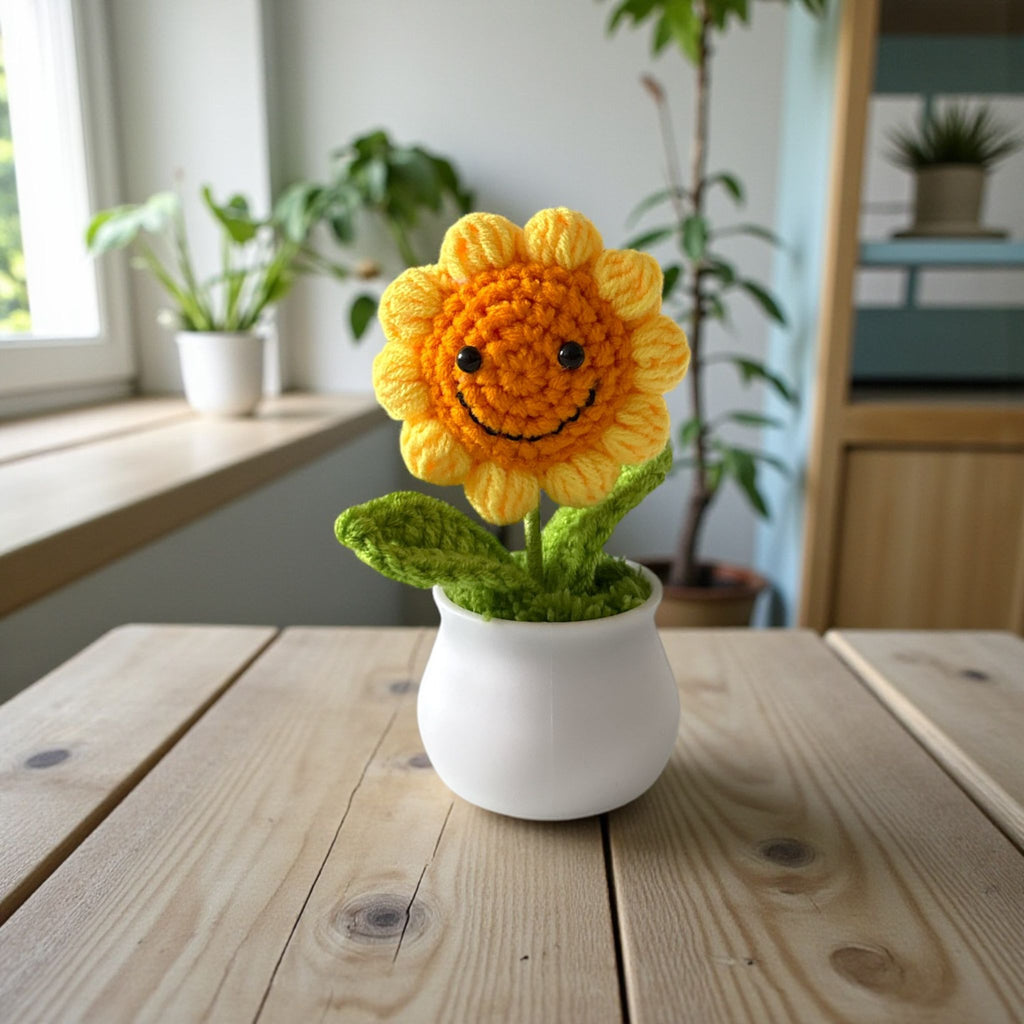 Crocheted sunflower in a white pot on a wooden table with plants in the background.