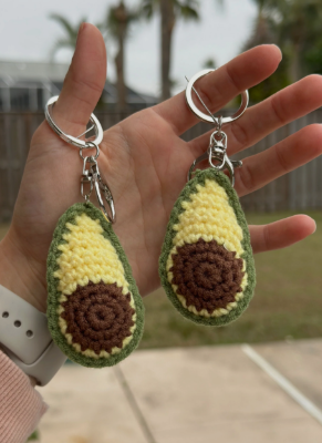 Hand holding two avocado-shaped keychains with a blurred outdoor background