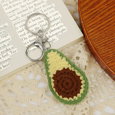 Crocheted avocado keychain on a textured surface with a book in the background