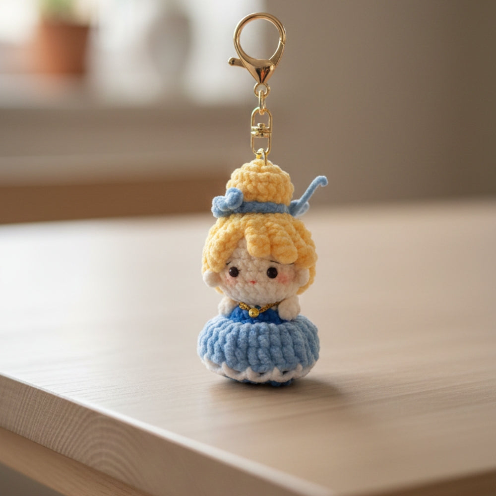 Crocheted keychain of a character with blonde hair and blue dress on a wooden surface.