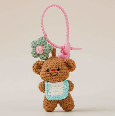 Crocheted teddy bear keychain with a flower and pink item on a beige background