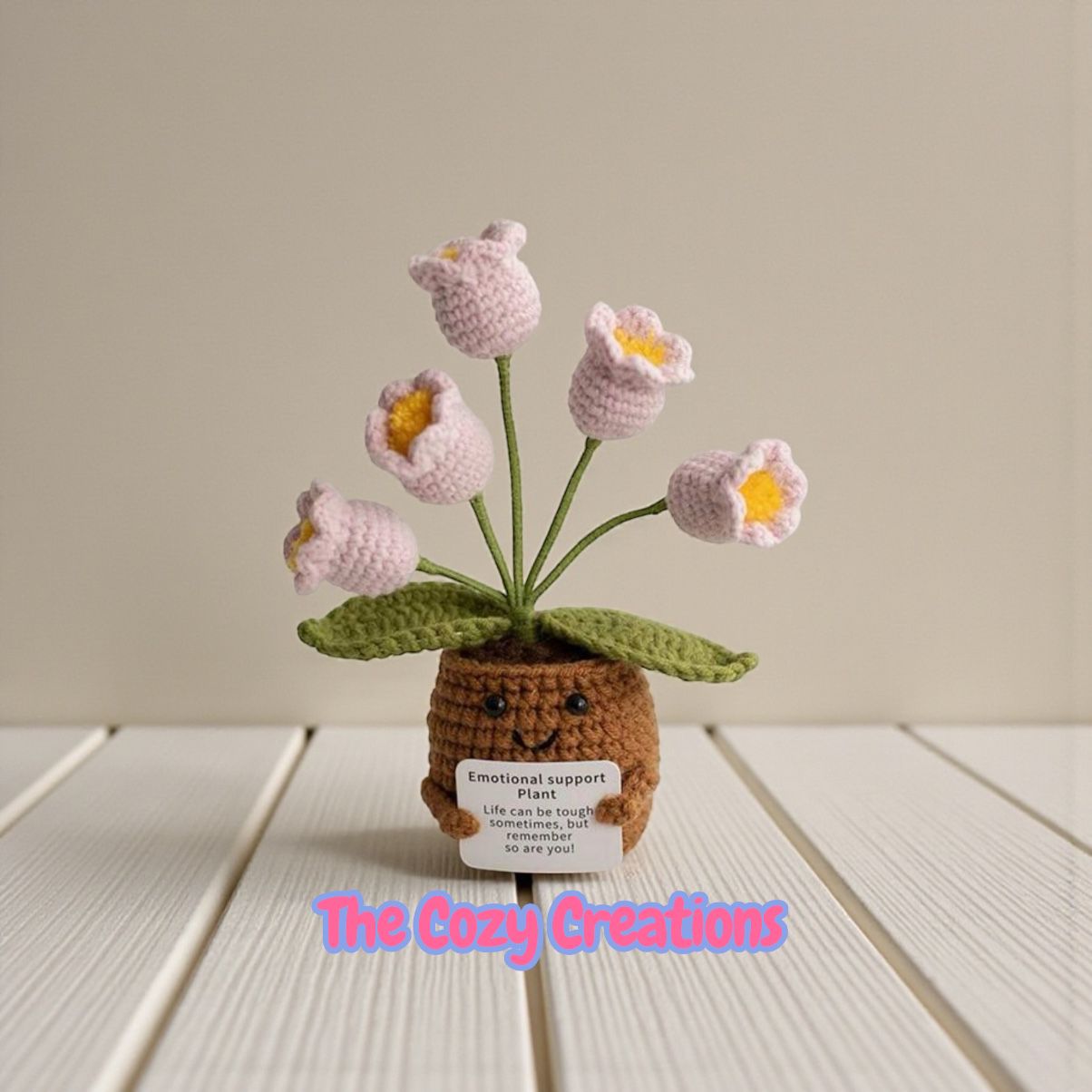 Crocheted flower plant with a face on a wooden surface, branded 'The Cozy Creations'.