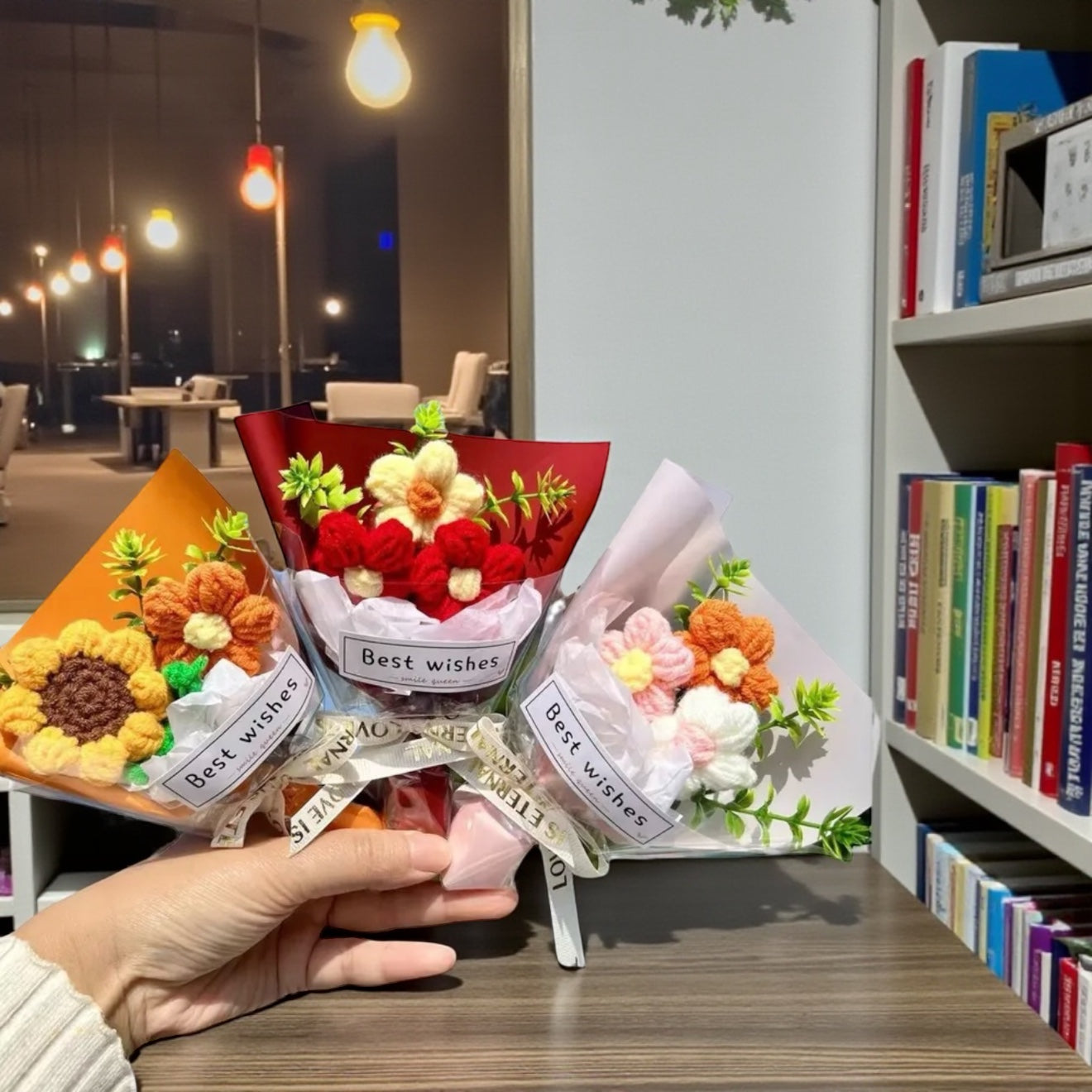 Three small bouquet packages labeled 'Best wishes' held by a hand in a room with books and decor.