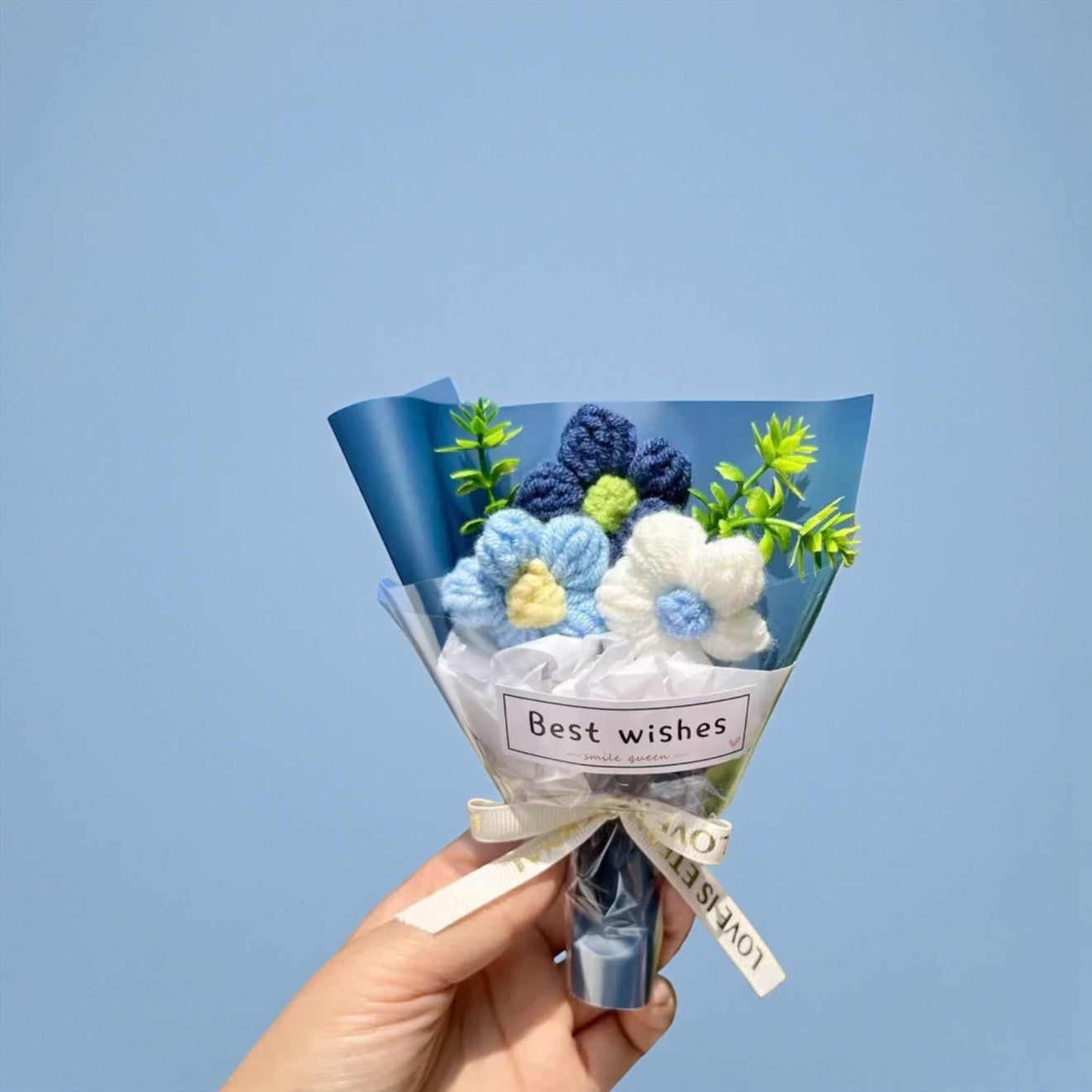 Hand holding a small bouquet of crocheted flowers with a 'Best wishes' tag against a light blue background