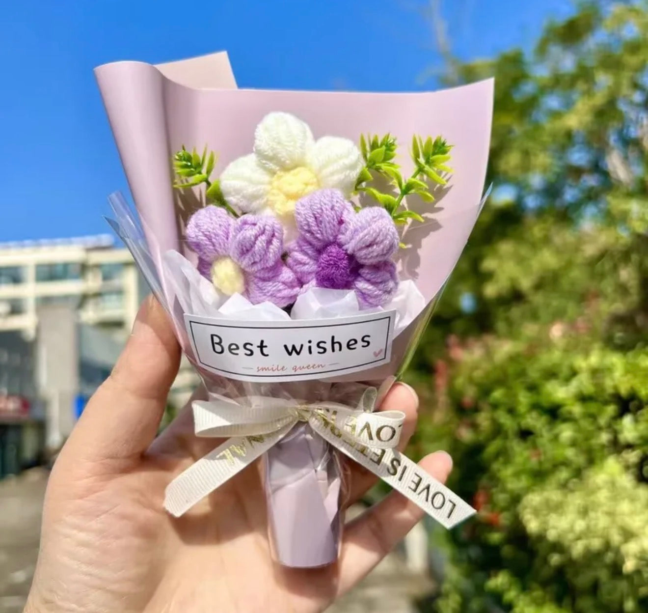 Bouquet of knitted flowers with a 'Best wishes' label held outdoors.