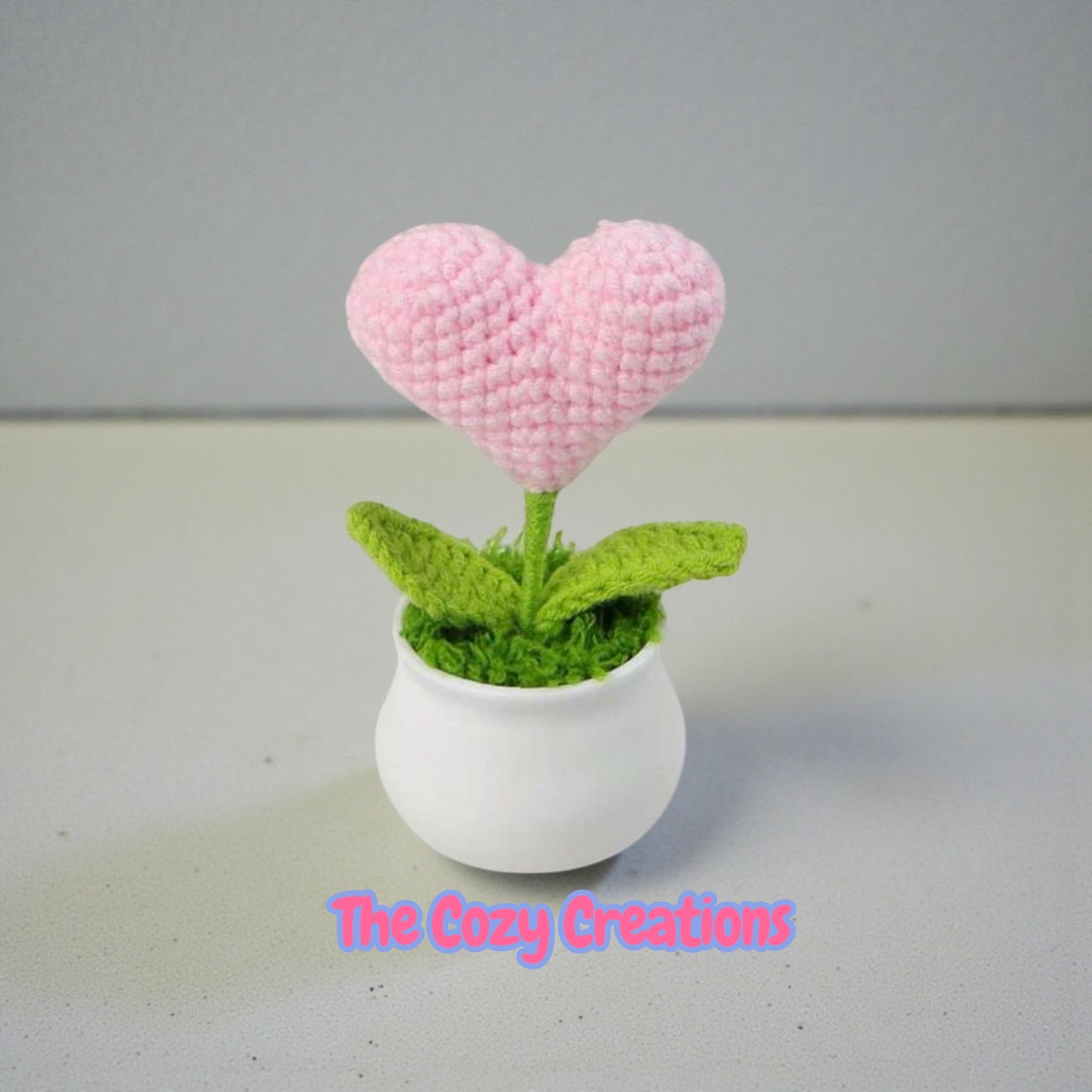 Heart-shaped pink flower in a white pot with green leaves on a gray background, branded 'The Cozy Creations'.