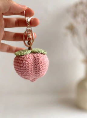 Pink crocheted strawberry keychain held by a hand against a neutral background