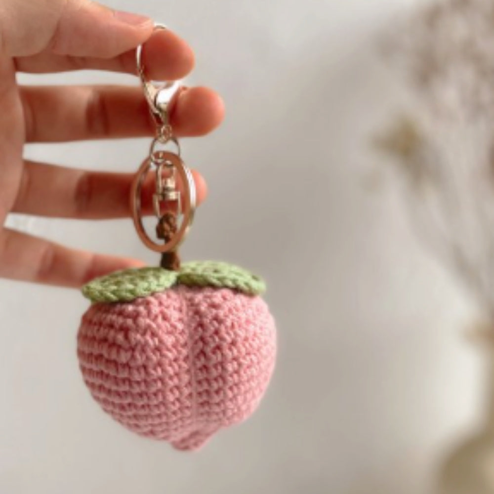 Hand holding a pink crocheted peach-shaped keychain with a green top.