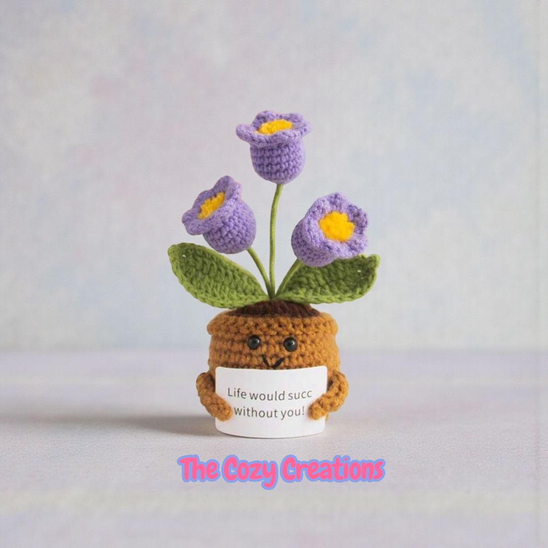 Crocheted flower pot with flowers and a small face, holding a card with text, on a light background.