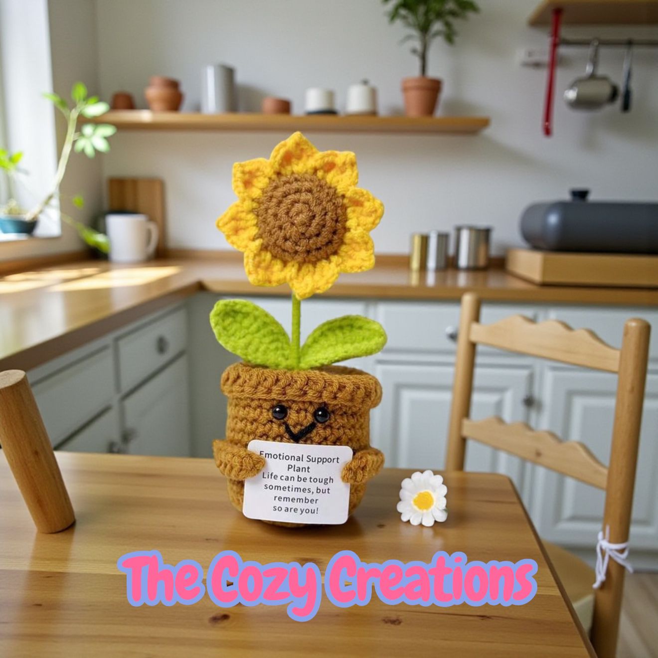 Crocheted sunflower plant with a face on a wooden table in a kitchen setting, branded 'The Cozy Creations'.