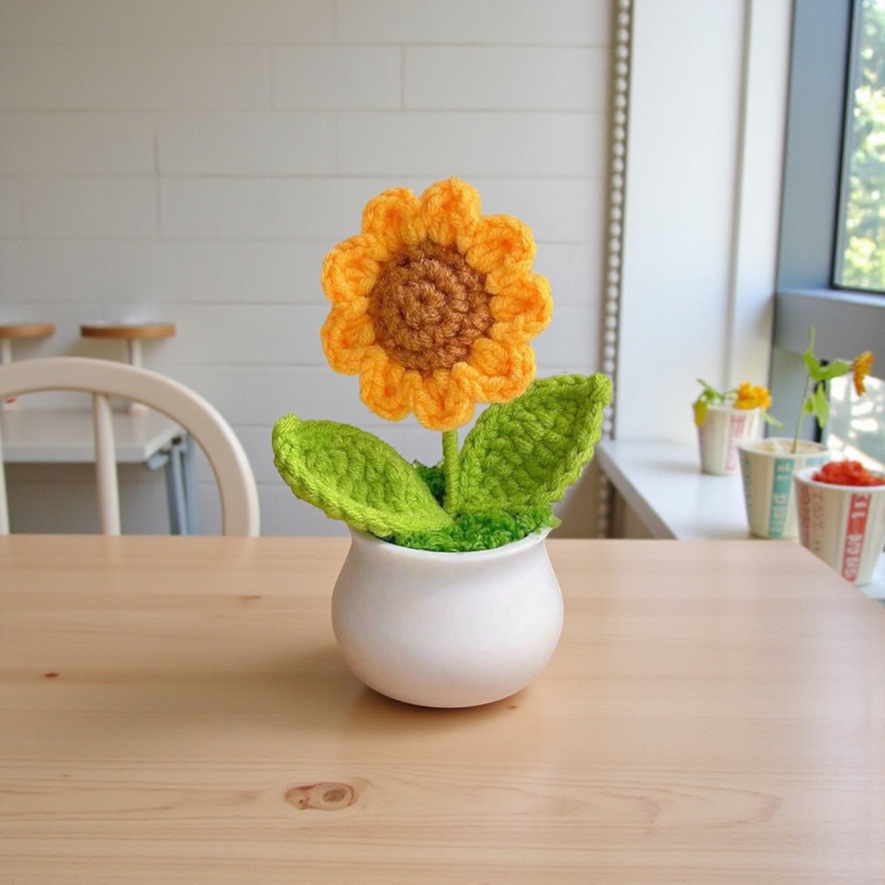 Crocheted sunflower in a white pot on a wooden table with a blurred indoor background.