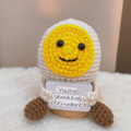Crocheted egg-shaped character with a yellow face and brown feet on a white surface.