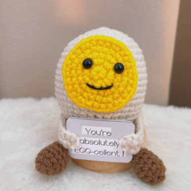 Crocheted egg-shaped character with a yellow face and brown feet on a white surface.