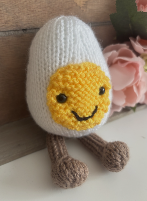 Knitted toy resembling a boiled egg with a smiling face on a wooden surface.
