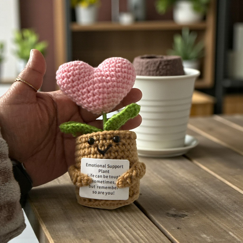 Hand holding a small crochet plant with a pink heart above it on a wooden surface.