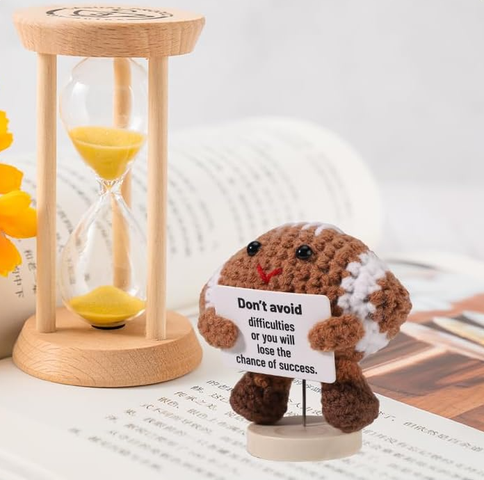 Small plush toy with a motivational message card next to a wooden hourglass on a surface.