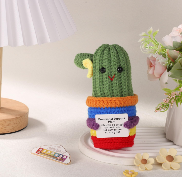 Colorful knitted cactus toy with a message tag on a white surface with decorative elements.