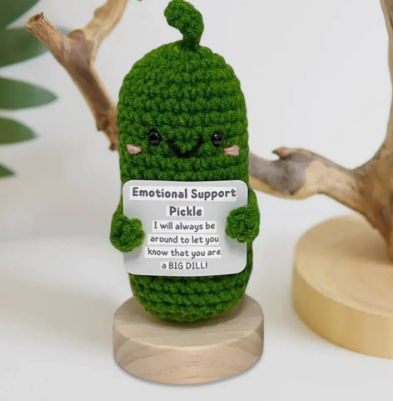 Crocheted pickle plushie holding a sign on a wooden stand with a white background