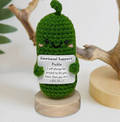 Crocheted pickle plushie holding a sign on a wooden stand with a white background