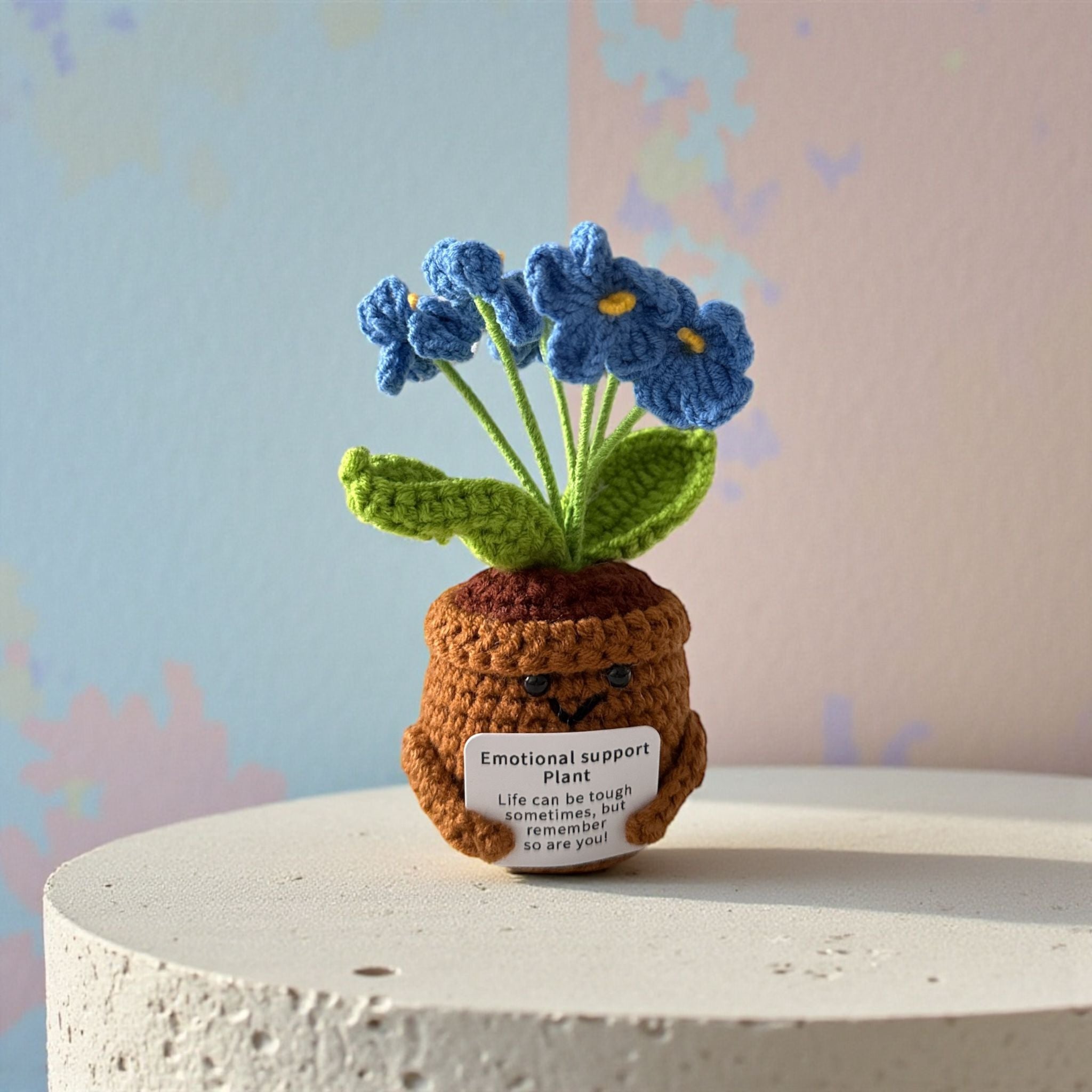 Crocheted emotional support plant with blue flowers on a white pedestal against a pastel background