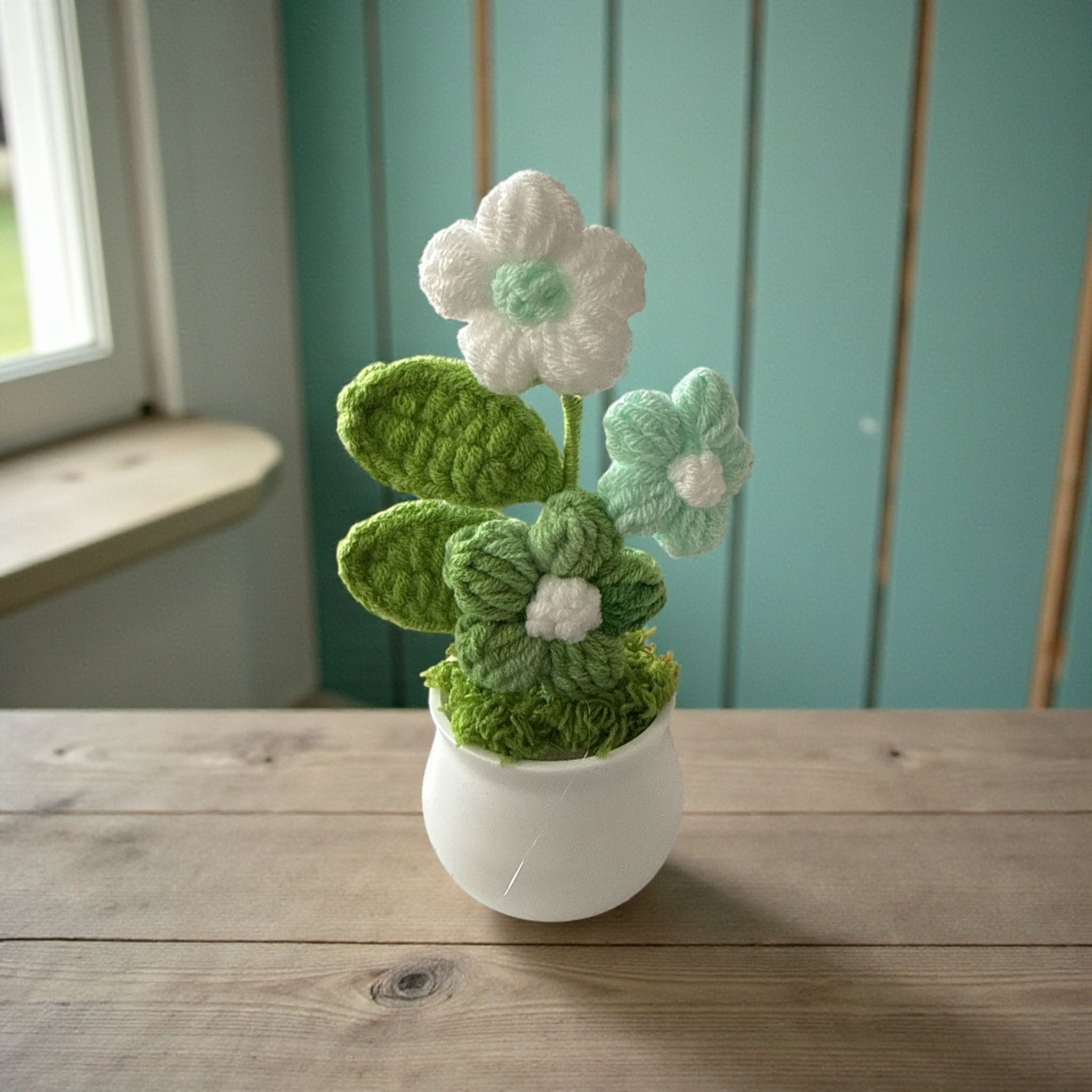 Knitted flower arrangement in a white pot on a wooden surface with a teal wall in the background.