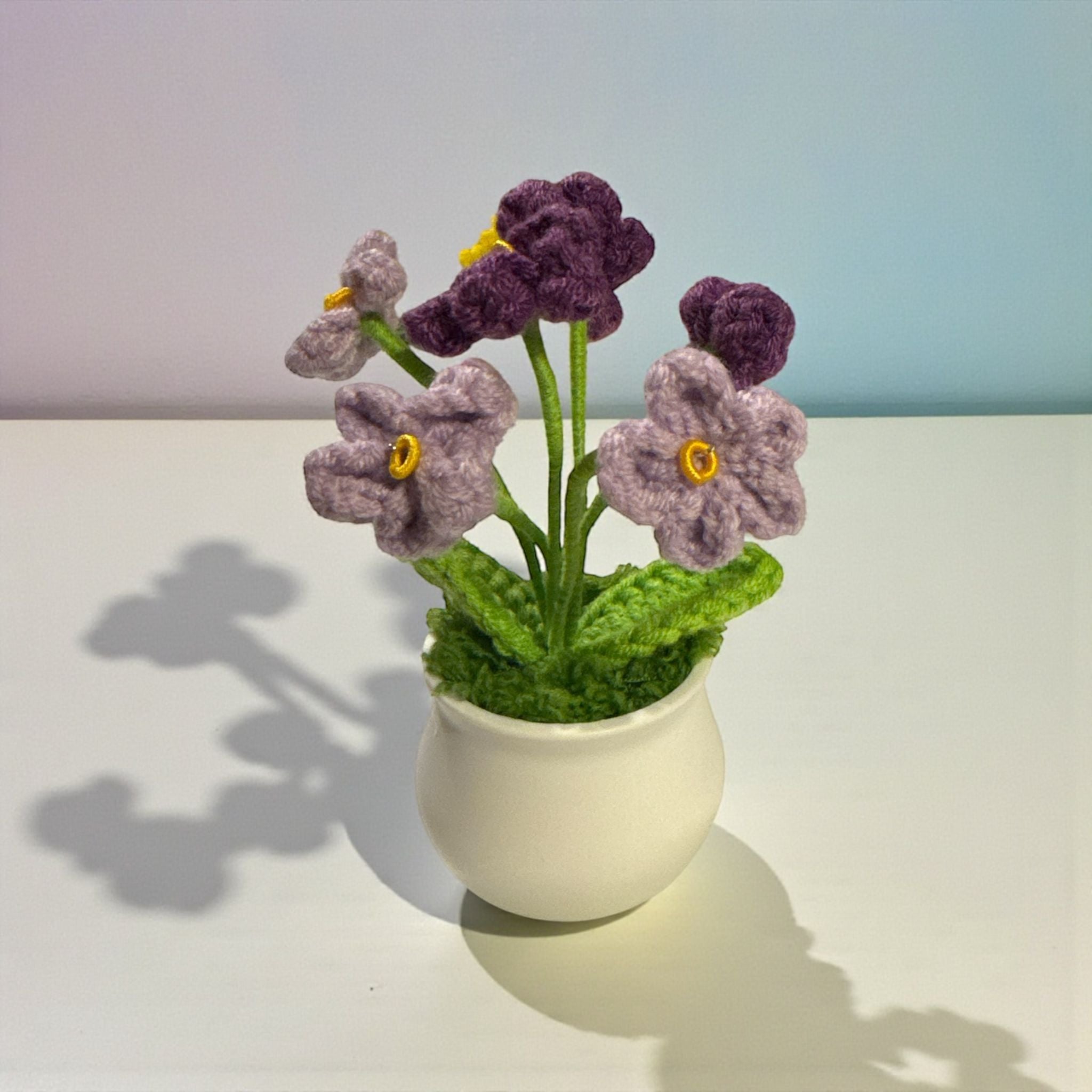 Knitted purple flowers in a white pot on a light surface with a blurred background