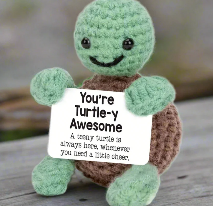 Crocheted turtle toy holding a sign saying 'You're Turtle-y Awesome' on a wooden surface.