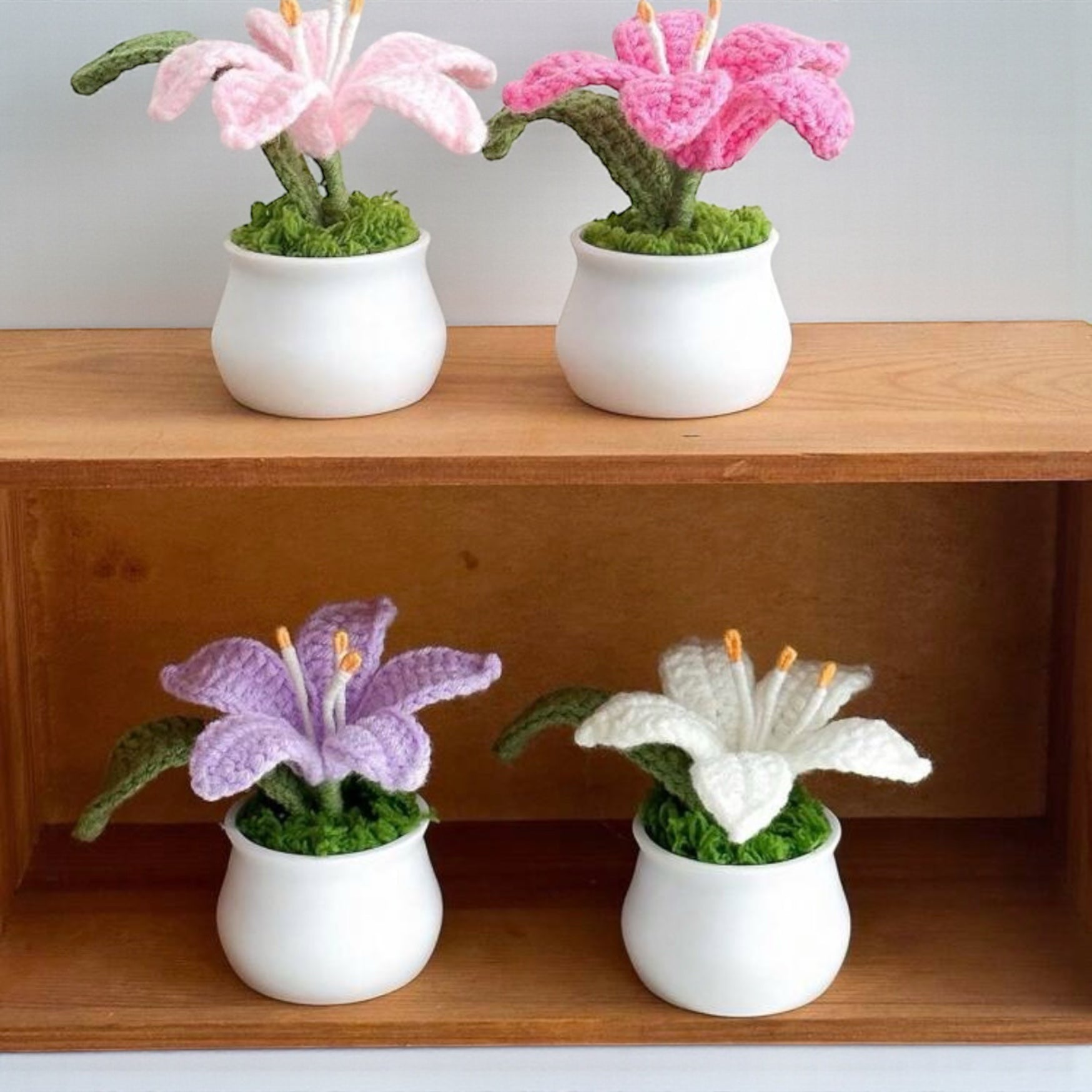 Four knitted flowers in white pots on a wooden shelf.