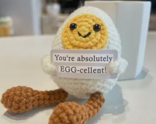 Crocheted egg-shaped character with a sign saying 'You're absolutely EGG-cellent!' on a white surface.