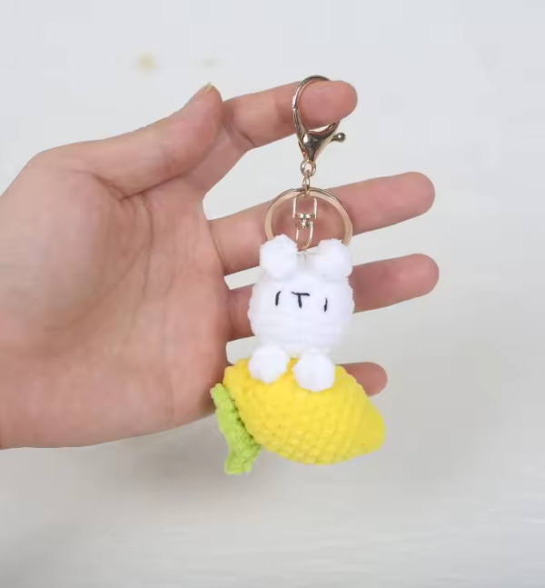 Hand holding a keychain with a white plush character on a yellow base against a light gray background