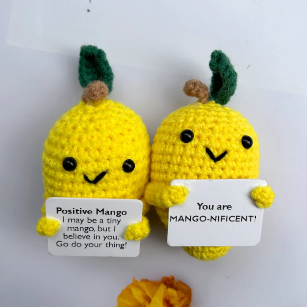 Two yellow crochet mangoes with smiling faces holding signs on a white background
