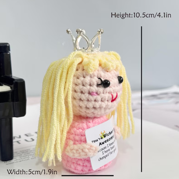 Small crochet doll with blonde hair and a pink dress, wearing a silver crown, on a light gray background.