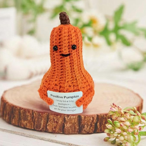 Crocheted pumpkin with a face on a wooden platform, surrounded by greenery.