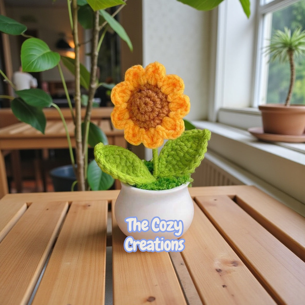 Crocheted sunflower in a pot on a wooden table with 'The Cozy Creations' branding.
