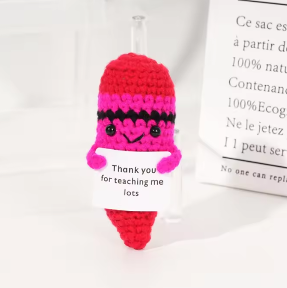 Crocheted character with a pink and red hat and black accents, sitting on a white surface with a text card.