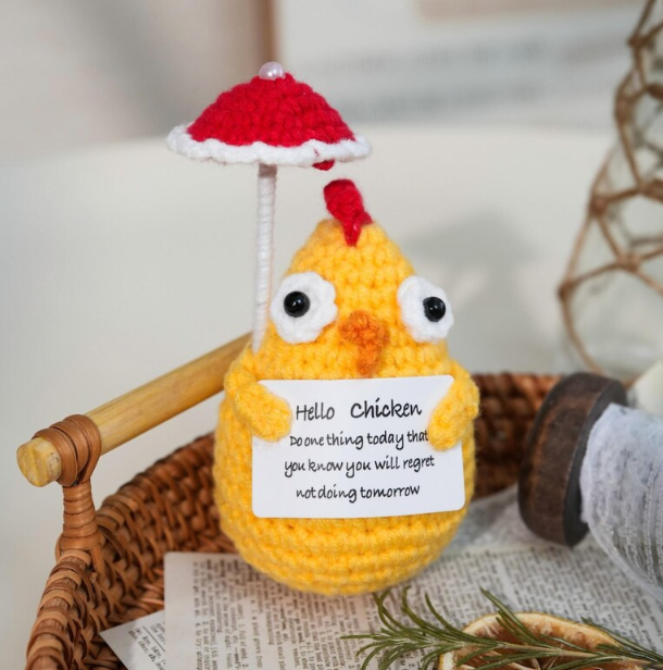 Crocheted yellow chicken with a red umbrella and humorous message on a woven surface.