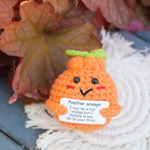 Crocheted orange character with a message tag on a textured surface with leaves in the background